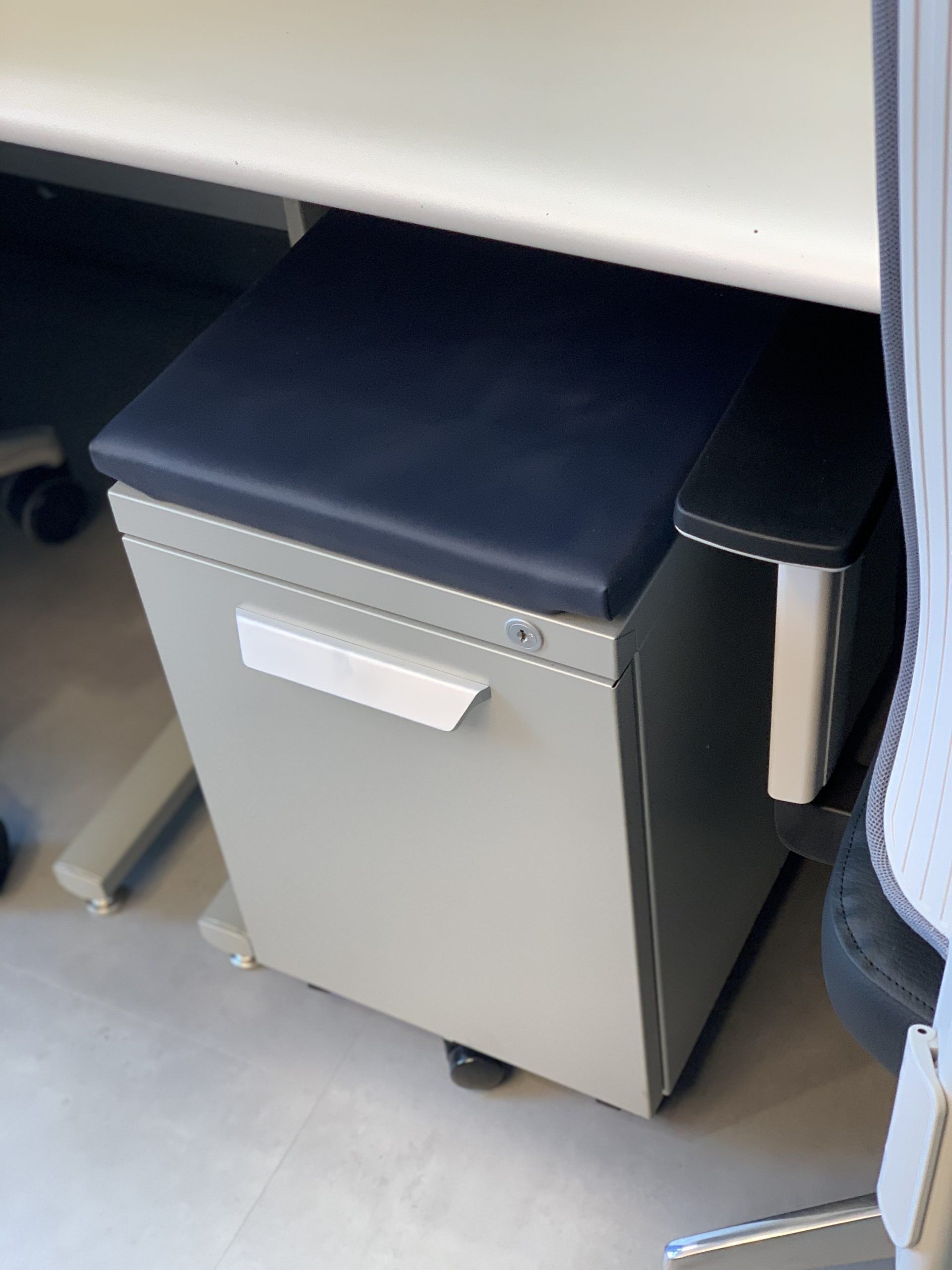 A filing cabinet under a desk with a black cushion on it.