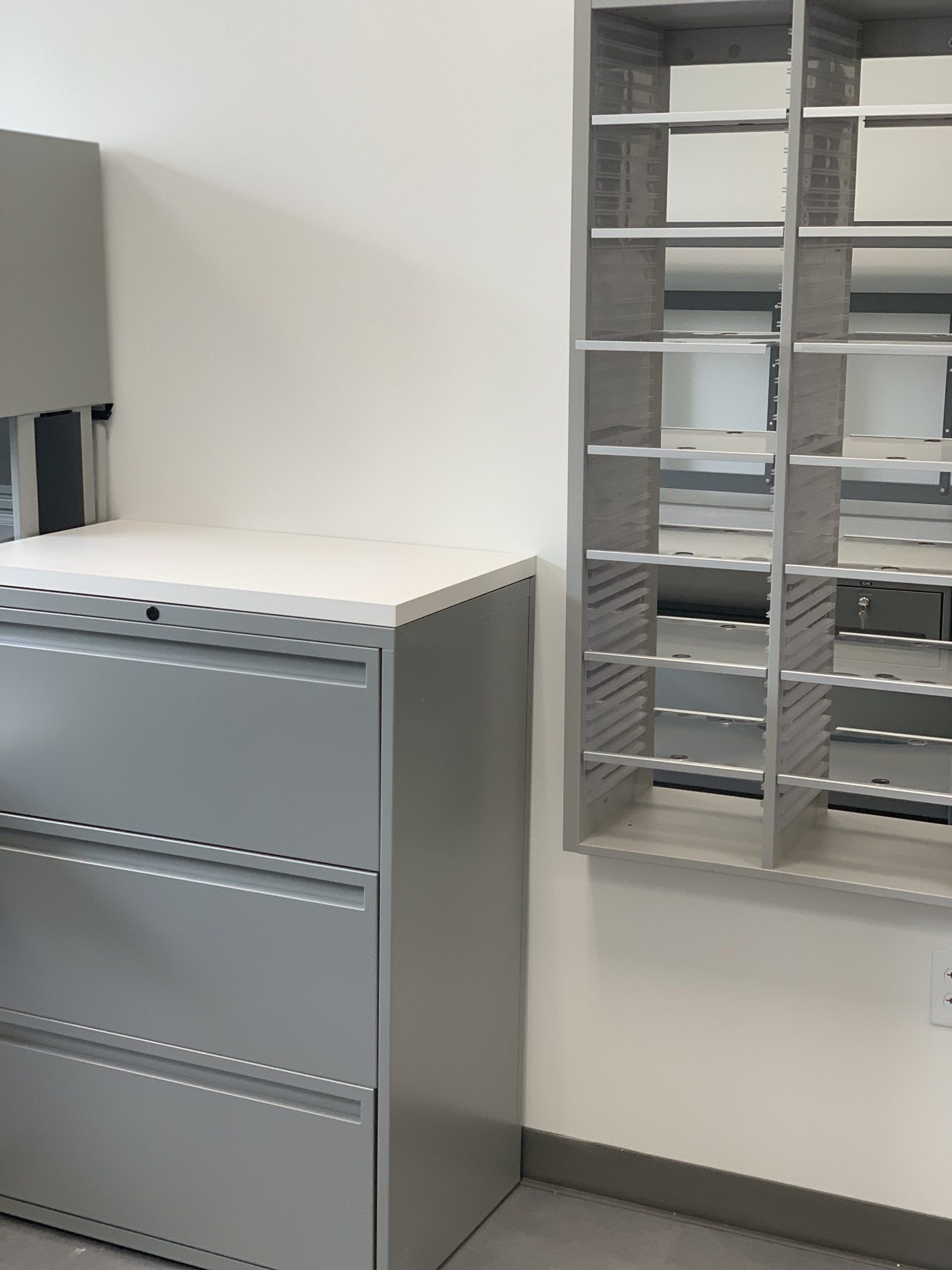 A gray filing cabinet sits next to a wall mounted shelf