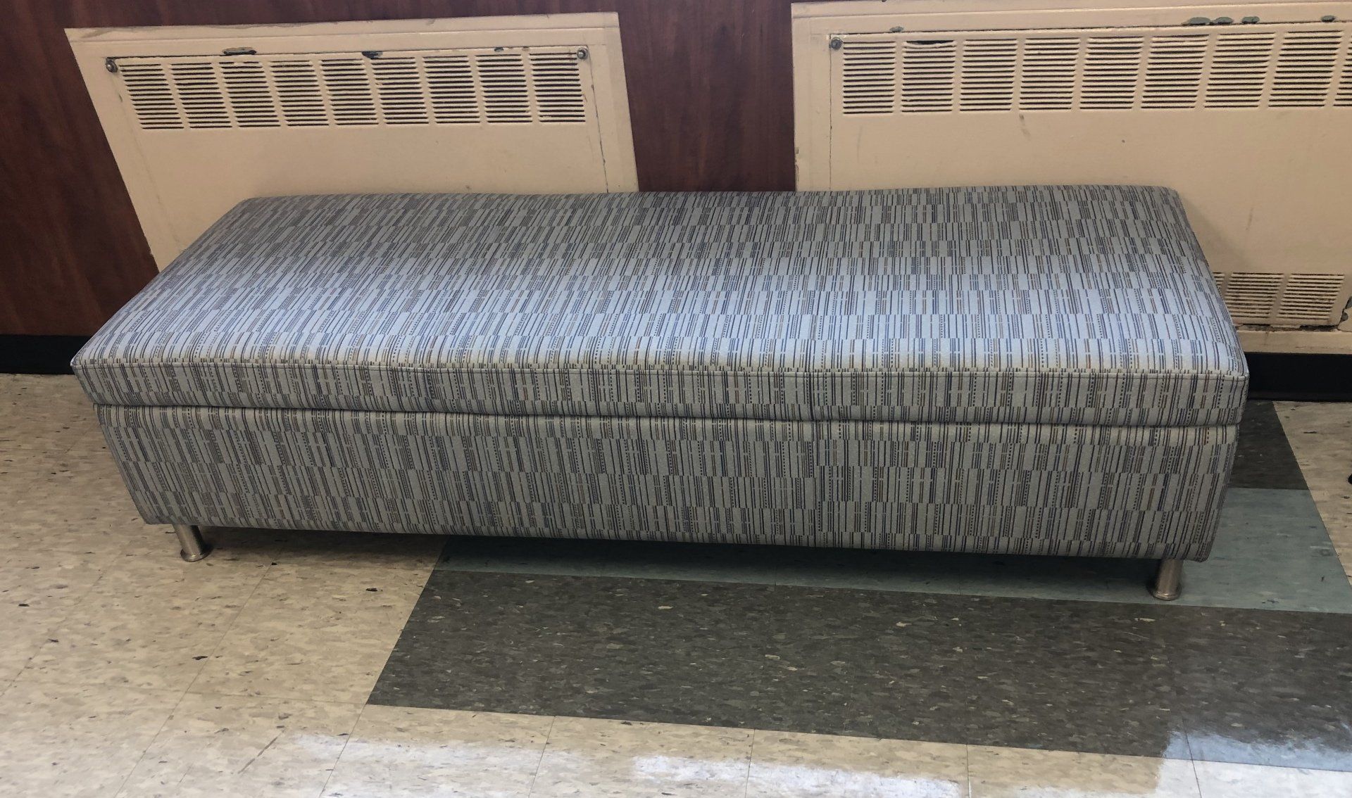 A gray ottoman is sitting on a tiled floor in a room.