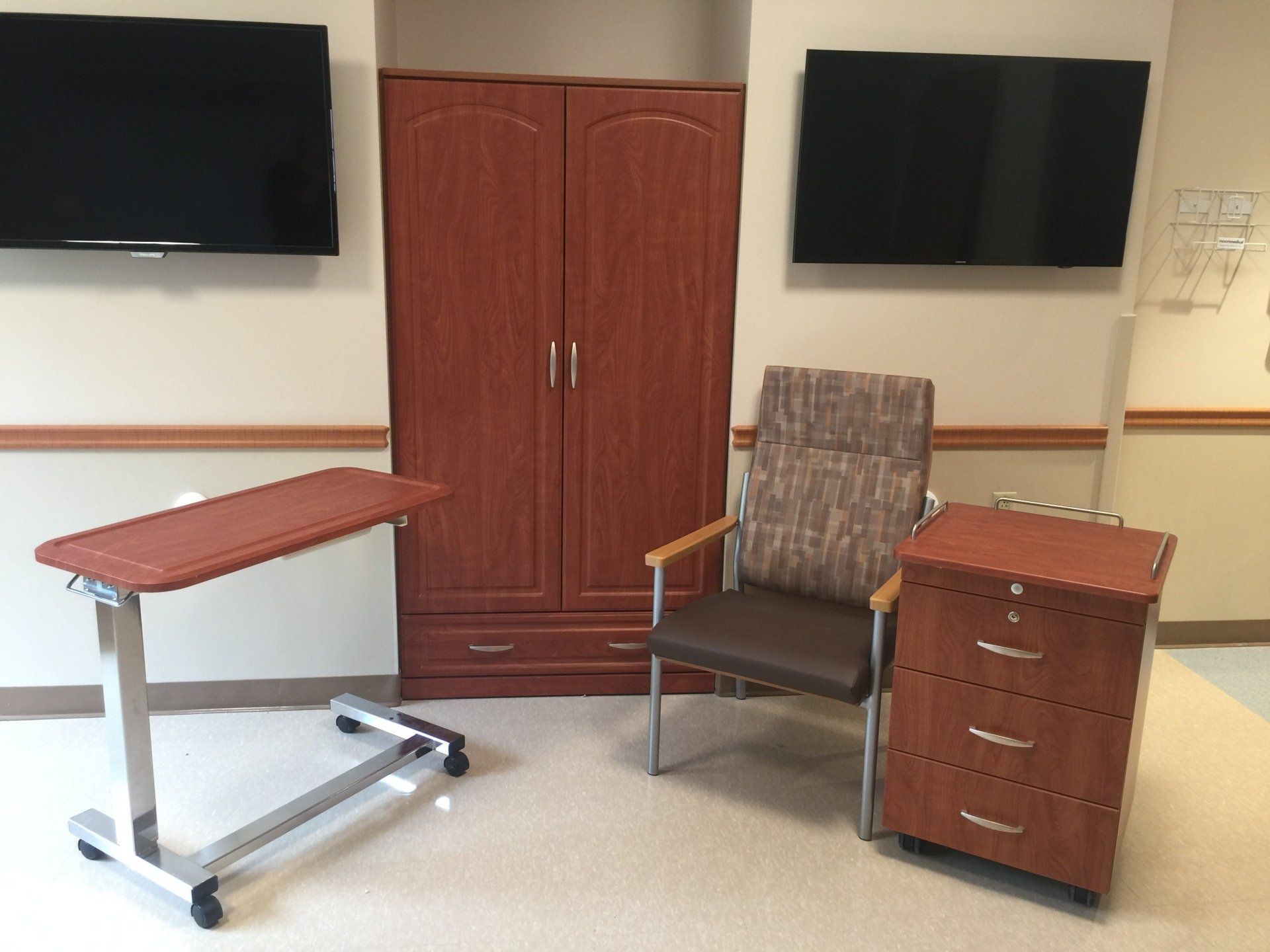 A hospital room with a chair a table and two televisions