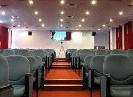 A large auditorium with rows of blue chairs and a tripod in the middle.