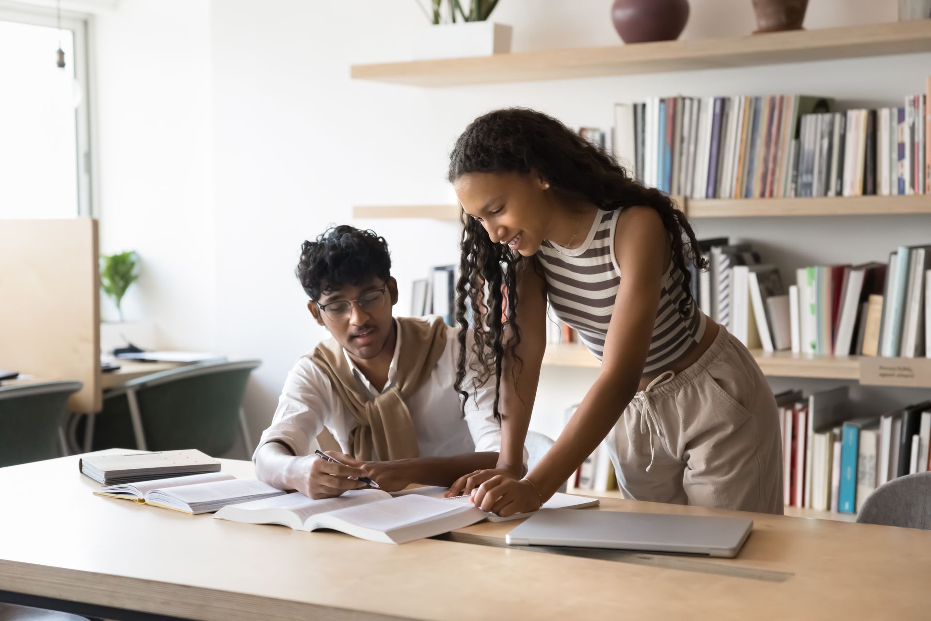 Two students studying together at a desk in a library, looking at a book and laptop.