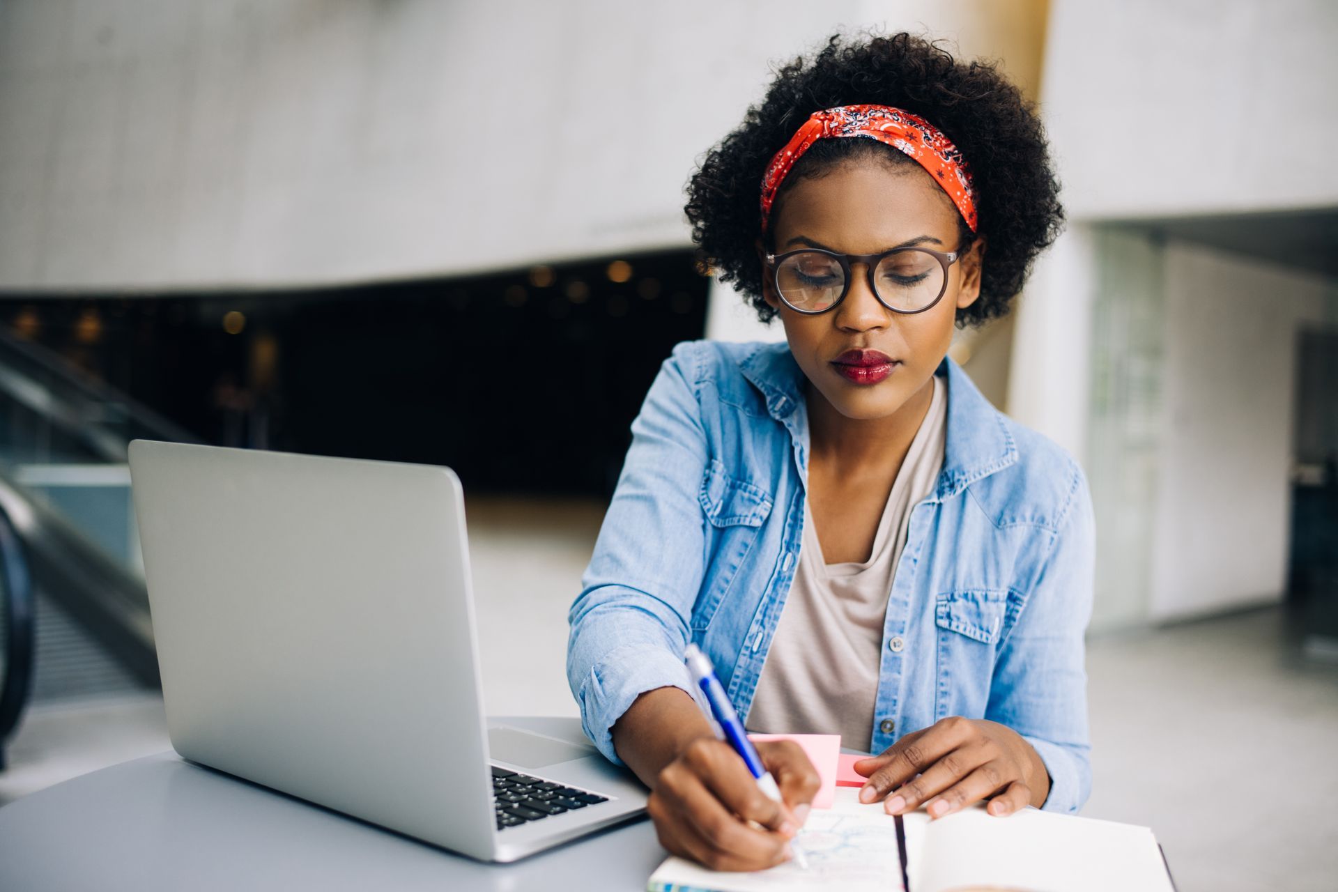 Woman with glasses writing in a notebook next to a laptop; outdoors with a light denim shirt, red headband.