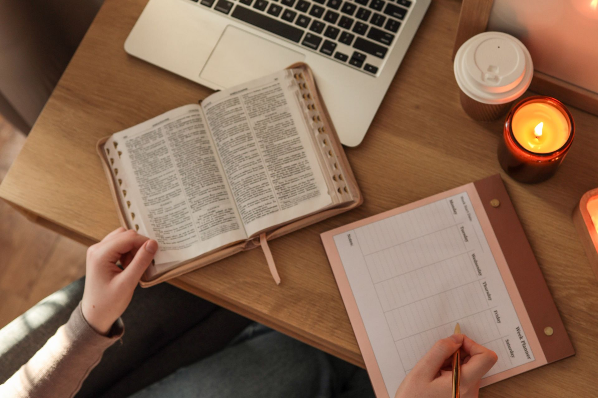 Hands holding an open book and writing on a notepad with a laptop, candle, and coffee cup on a wooden desk.