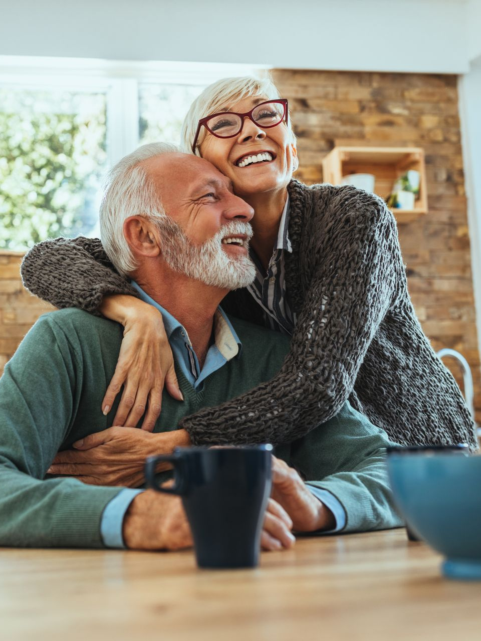 Smiling couple embracing at a table, woman hugging man from behind, coffee cup and bowl visible.