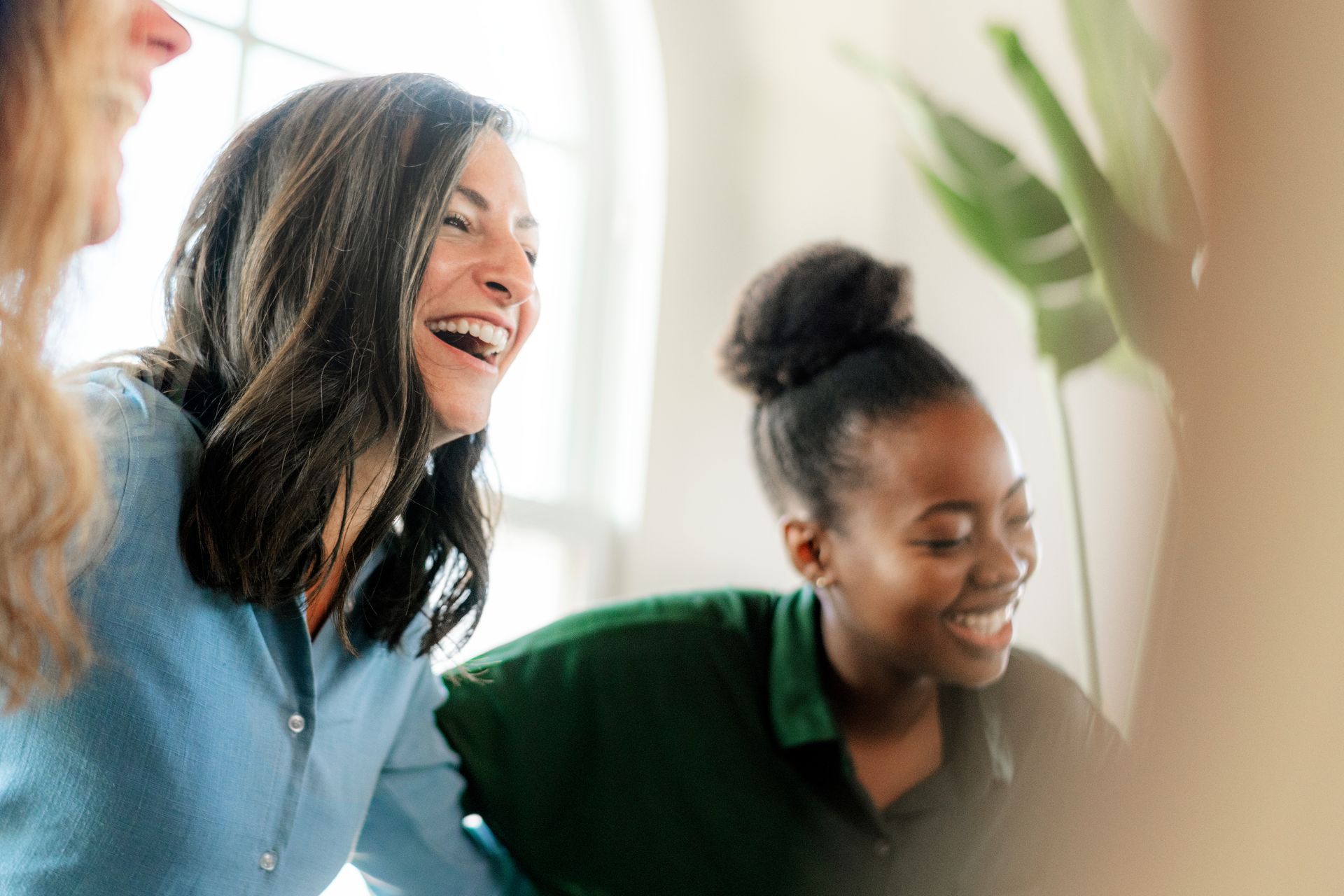 Three women laugh, one with dark hair in a bun. Bright, indoors setting with greenery.
