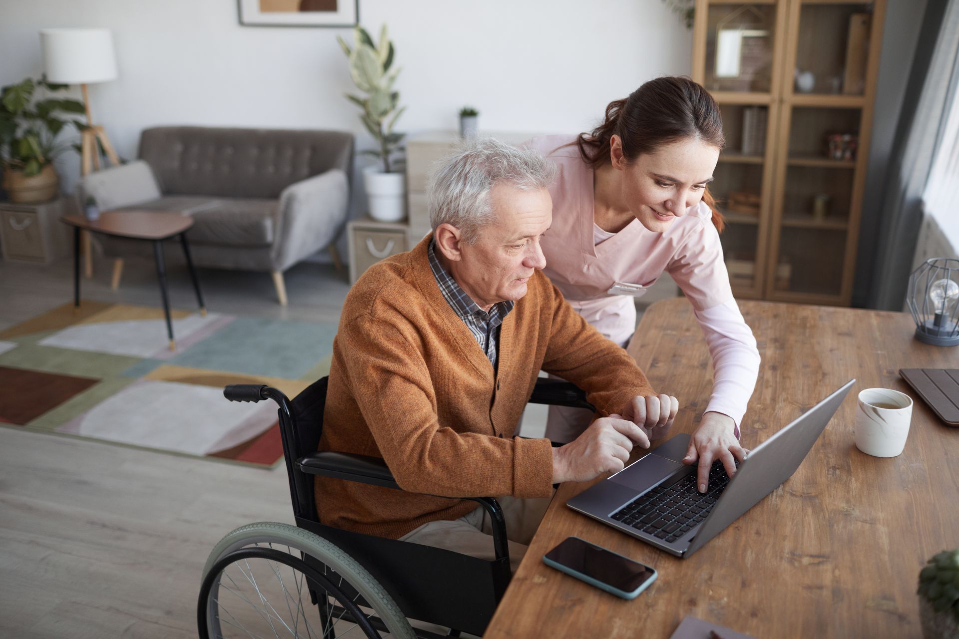 Woman assisting a person in a wheelchair with a laptop at a wooden table.