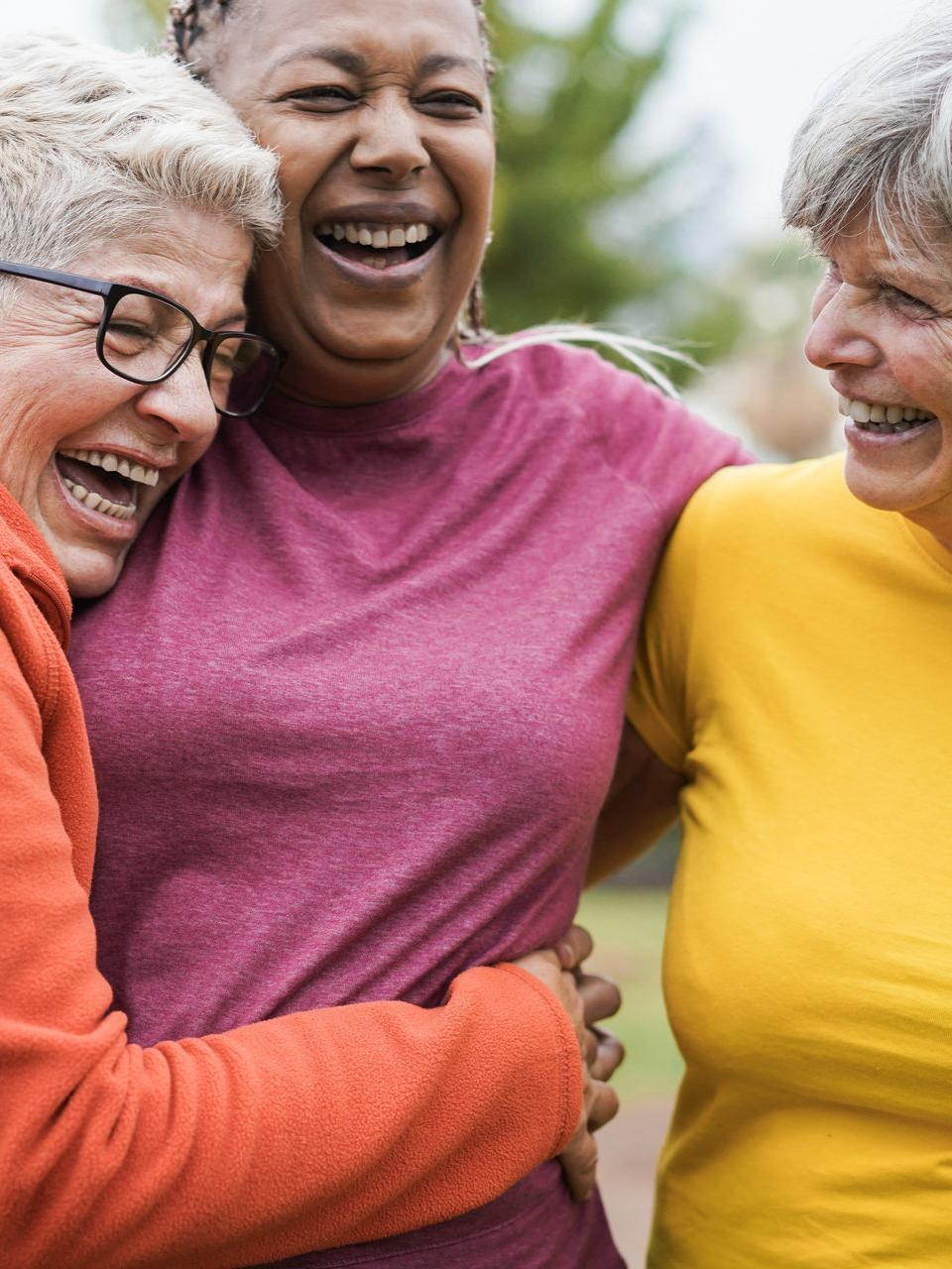 Three smiling women embracing outdoors. One wears orange, pink, and yellow shirts.