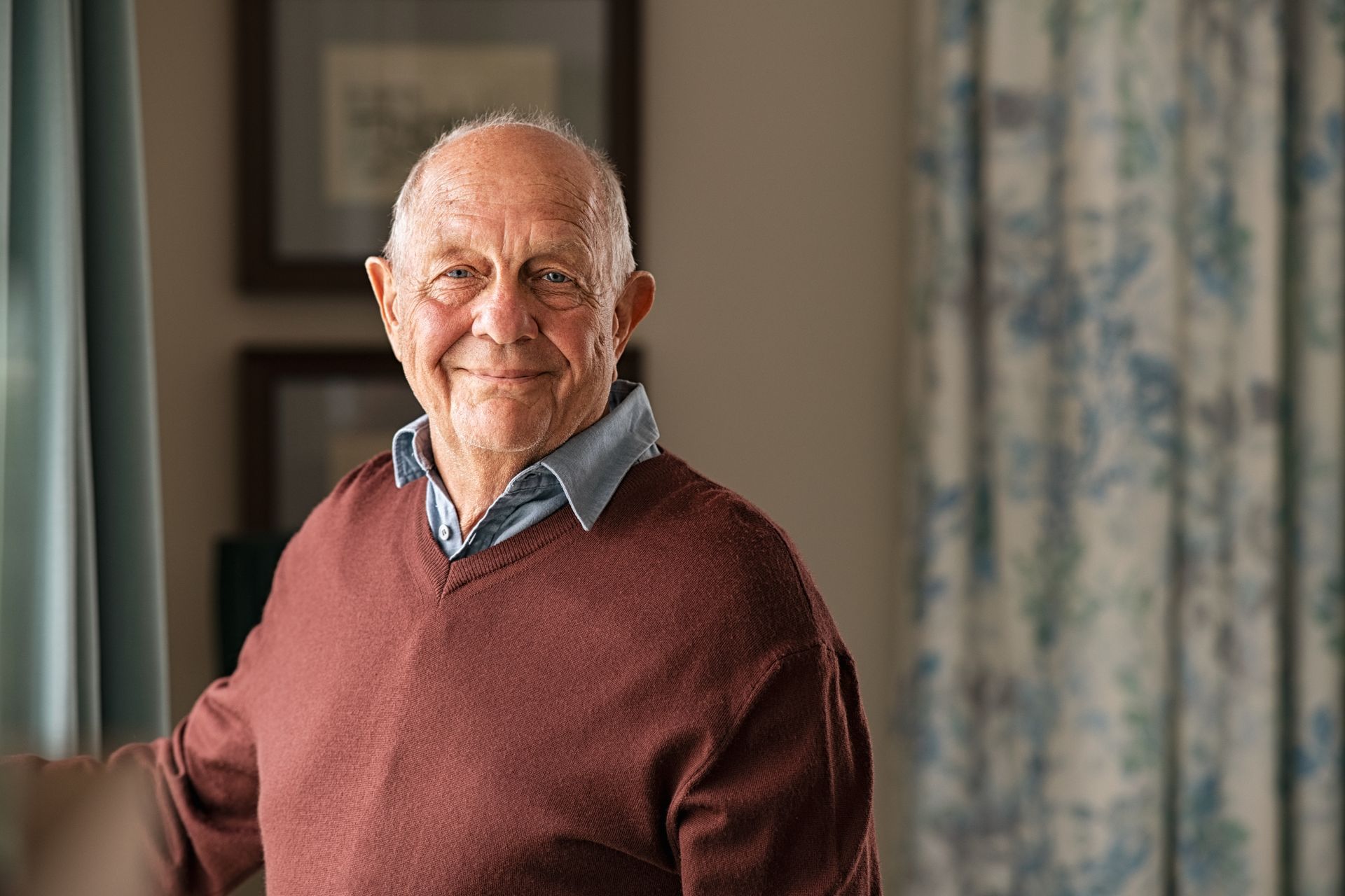 Older man wearing a red sweater smiles, leaning by a window, near a patterned curtain.