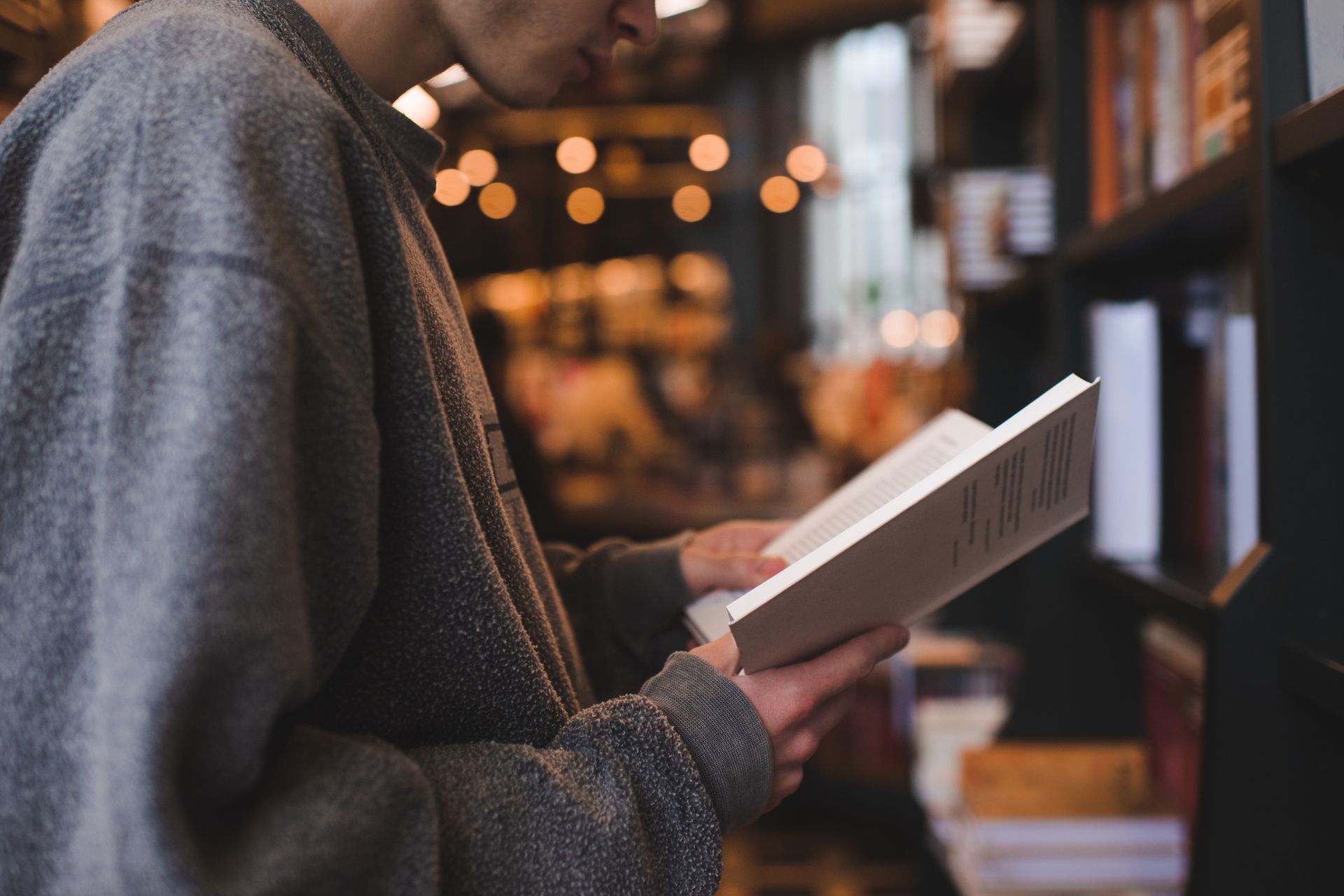 Person in gray sweater reads a book in a library, shelves in background.