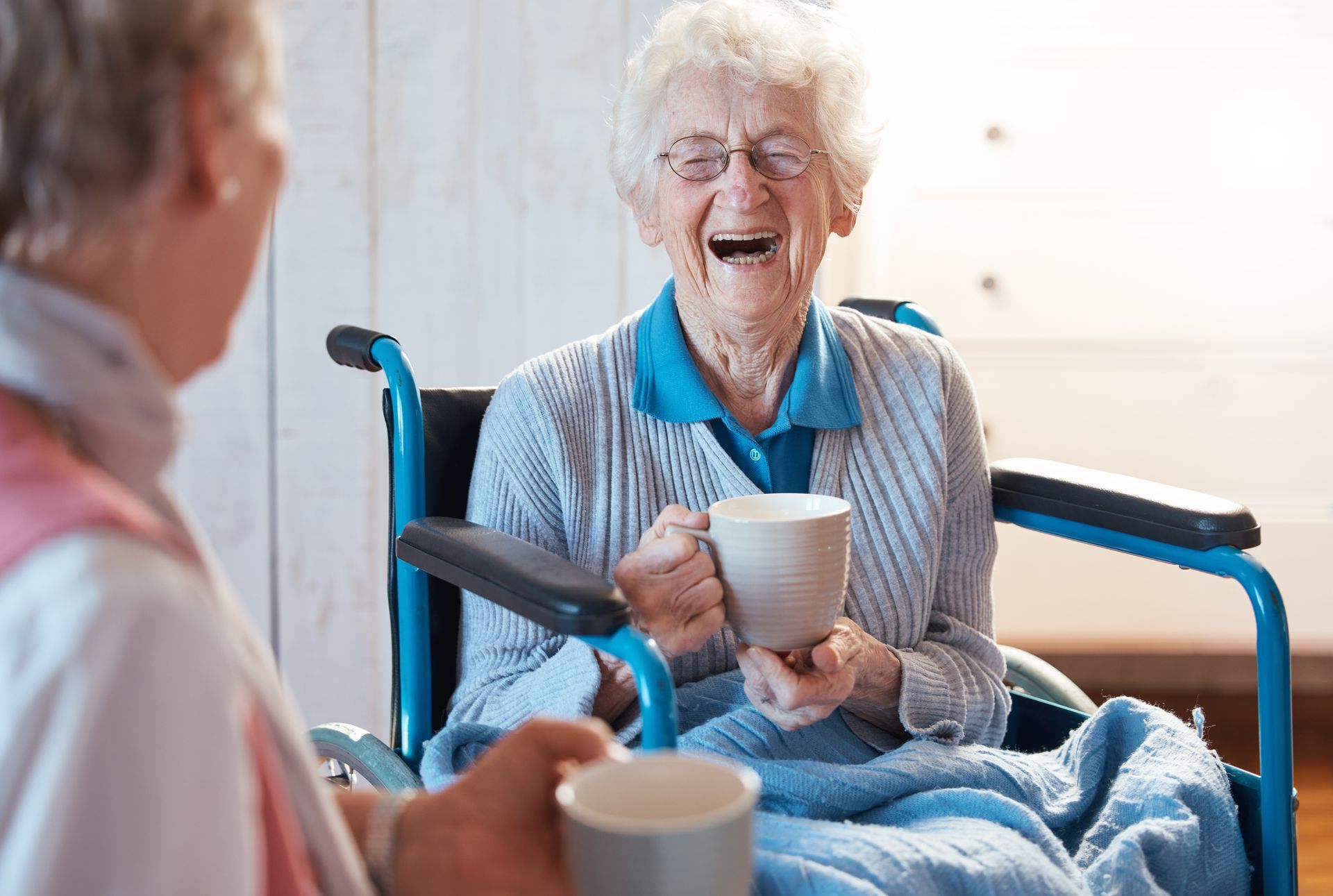 Woman in wheelchair laughs while holding a mug, conversing with another person holding a mug. Indoor setting.