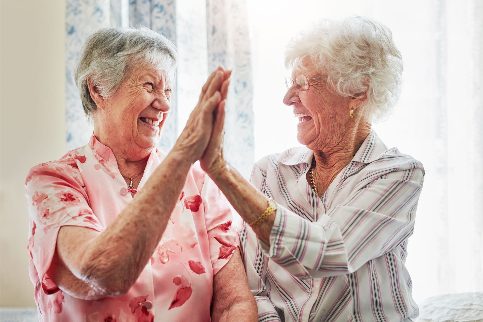 Two elderly women high-fiving, smiling. One wears pink, the other a white-striped shirt, indoor setting.