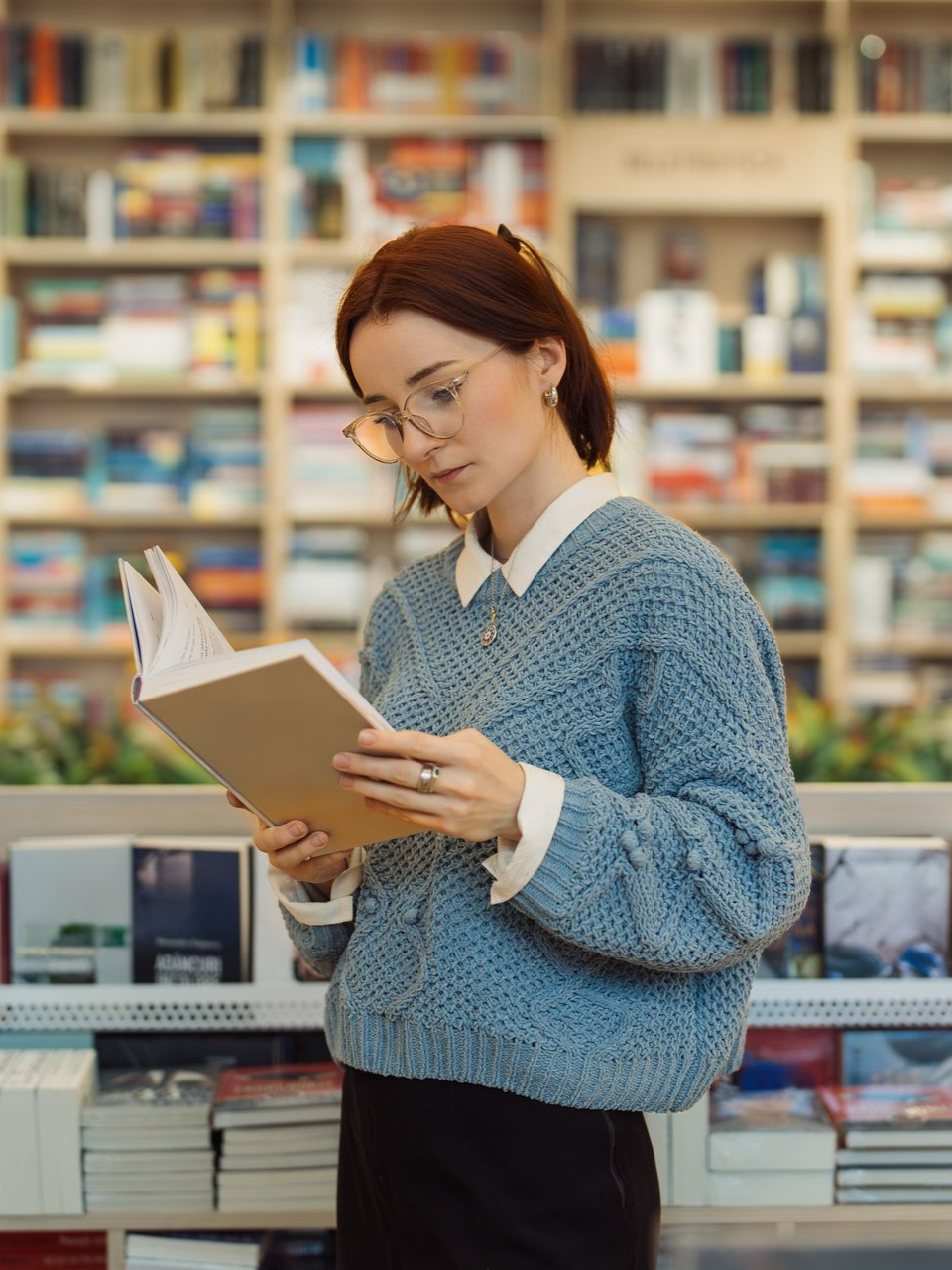 Woman in glasses reads a book in a bookstore, shelves of books in the background.
