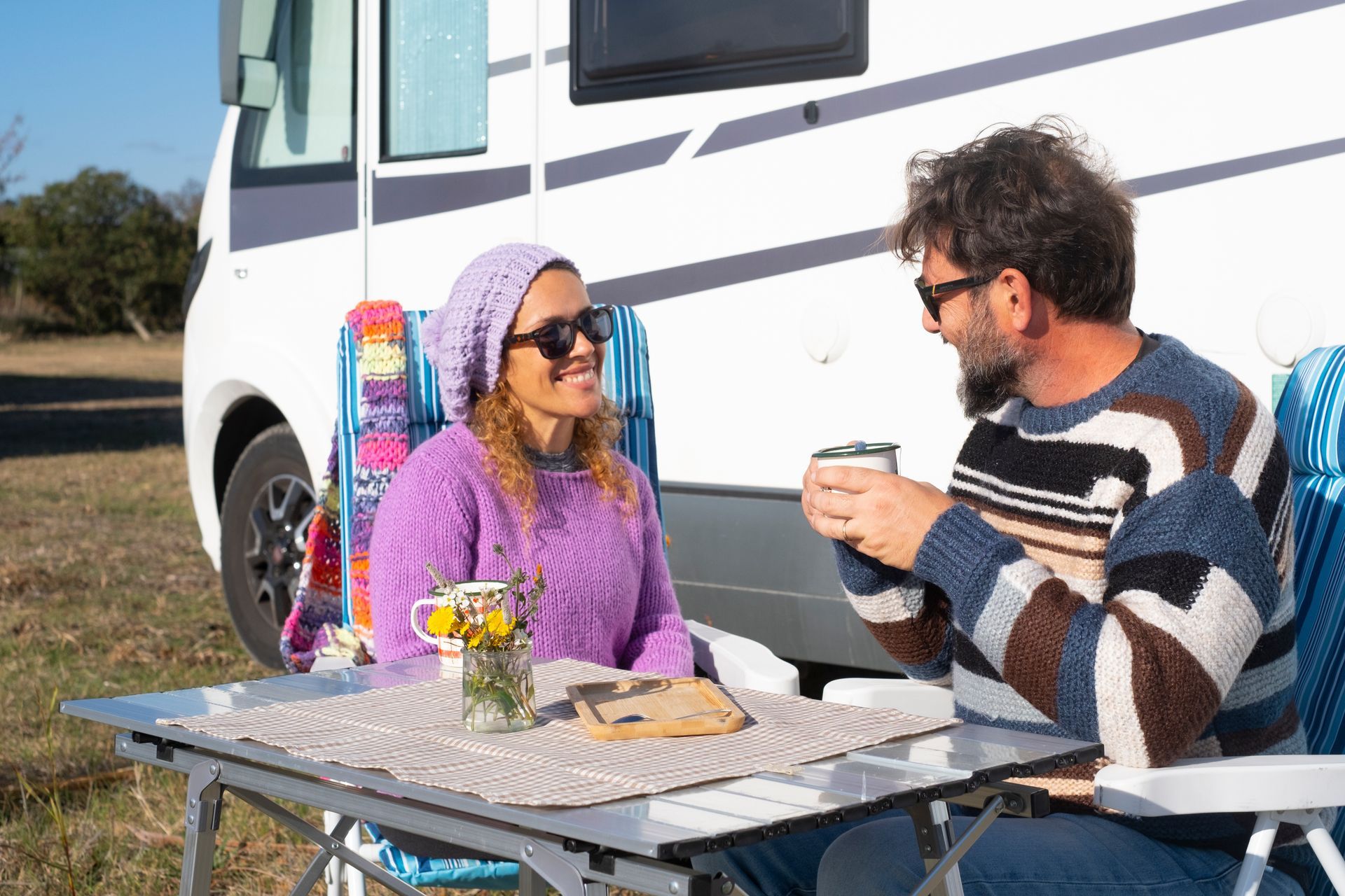 A couple in knit sweaters sits at a folding table outside a camper van, smiling and drinking from mugs on a sunny day.