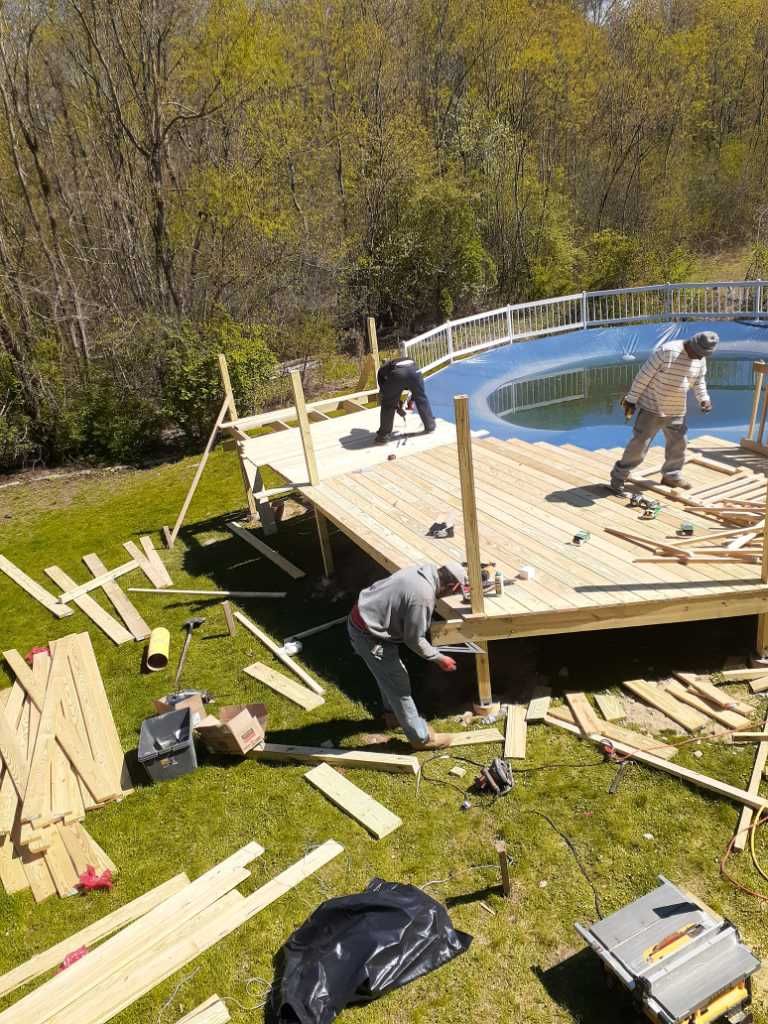Workers building a wooden deck around an above-ground swimming pool in a grassy backyard.