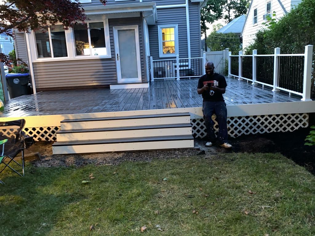 Man on deck holding phone, facing camera. Deck with steps, lattice, and white railing. Green grass in front of house.