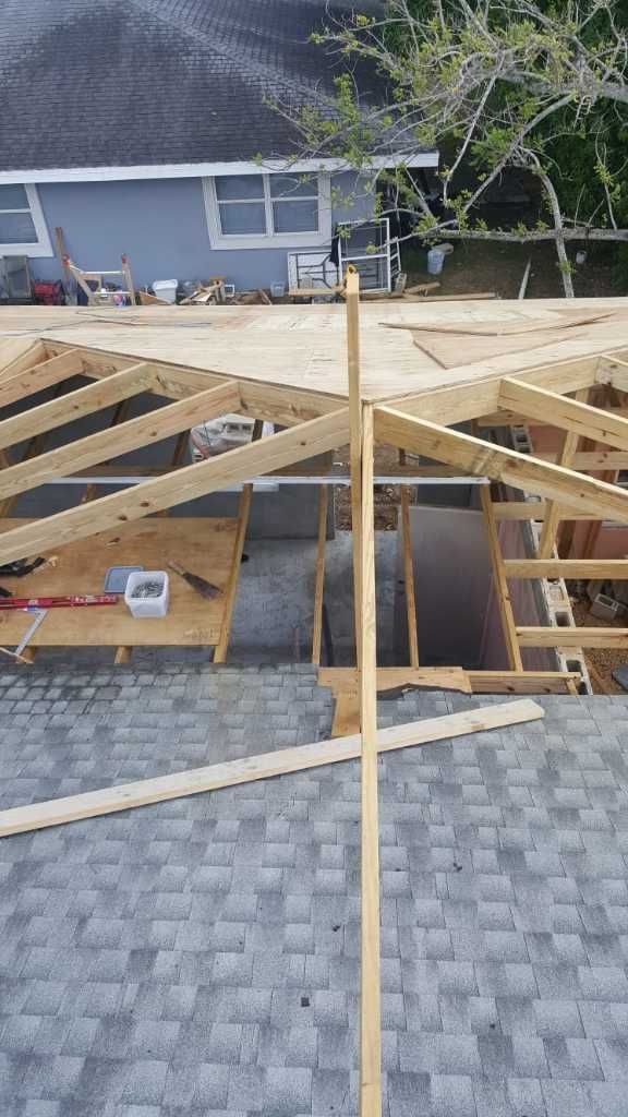Roof construction with exposed wooden trusses and sheathing, gray shingles, and a light blue house in the background.