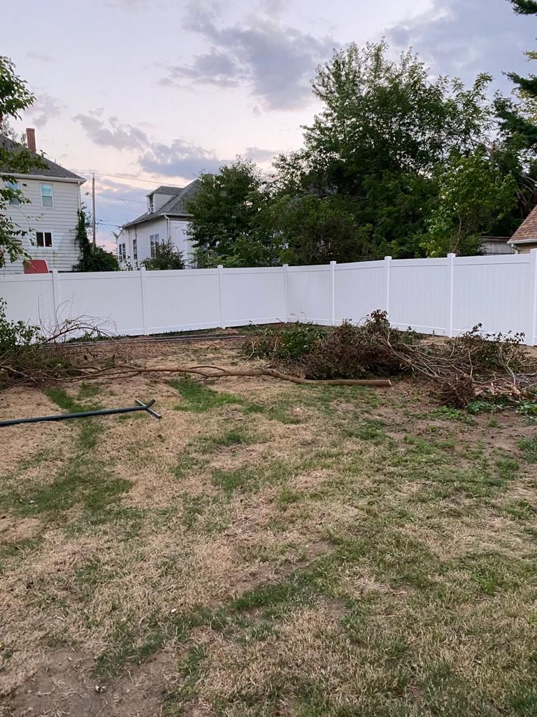 A white fence surrounds a backyard with grass and scattered brush; houses and trees in the distance.