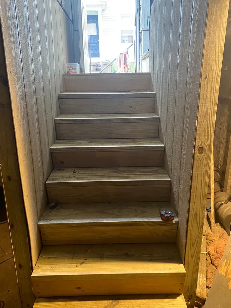 Wooden stairs leading up to a door, viewed from below. Light streams in from the top, illuminating the steps.