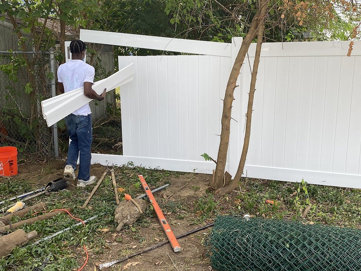 Man in jeans and a white shirt installing white fence panels outdoors.