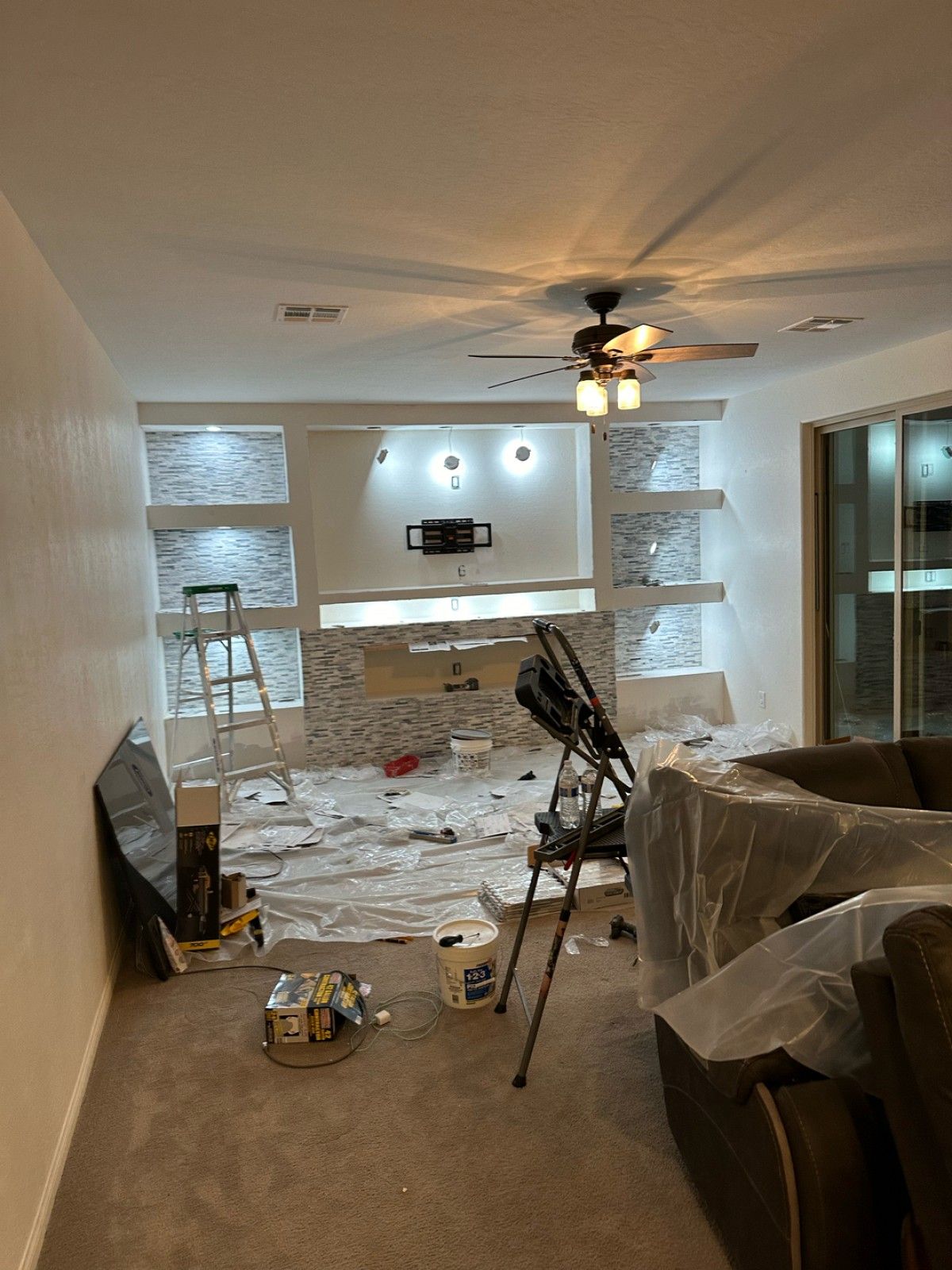 Living room renovation in progress, featuring built-in shelves with textured stone, ladder, and protective coverings on furniture.