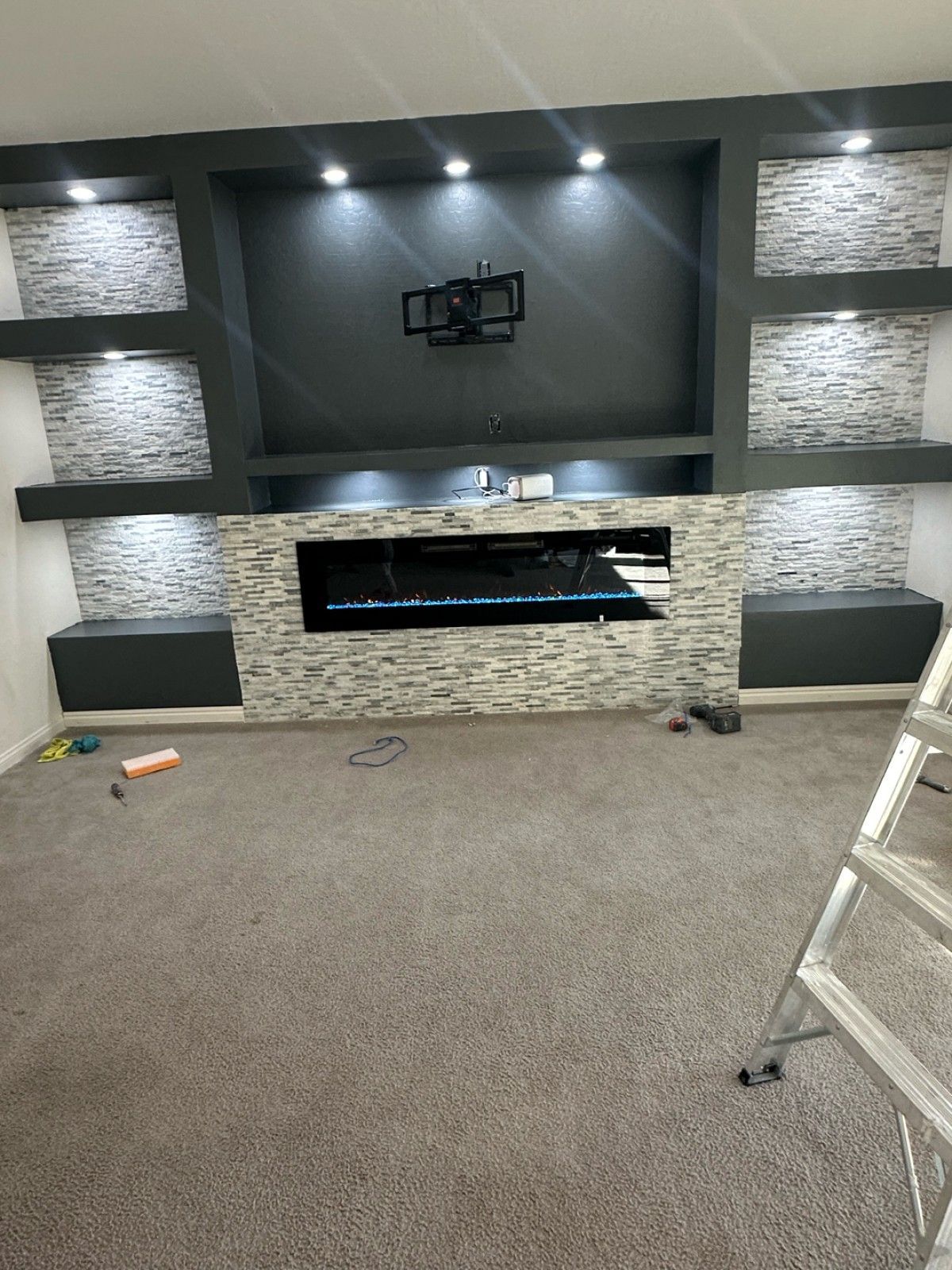 Gray entertainment center with stone accent, fireplace, and built-in shelves. Mounted TV, recessed lighting, on gray carpet.