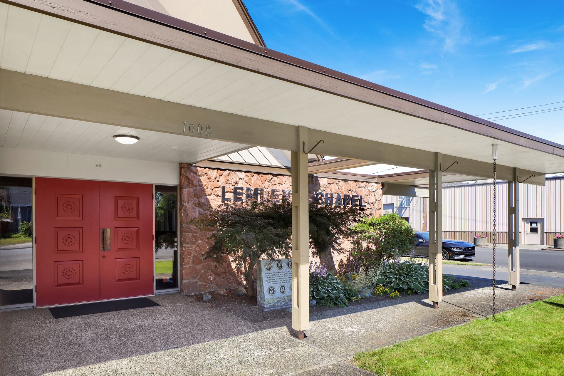 Building entrance with red double doors, covered walkway, and white support columns beside a grassy area.