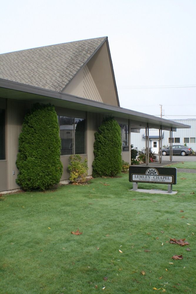 Small building with covered entrance, trimmed shrubs, and a lawn sign on a cloudy day