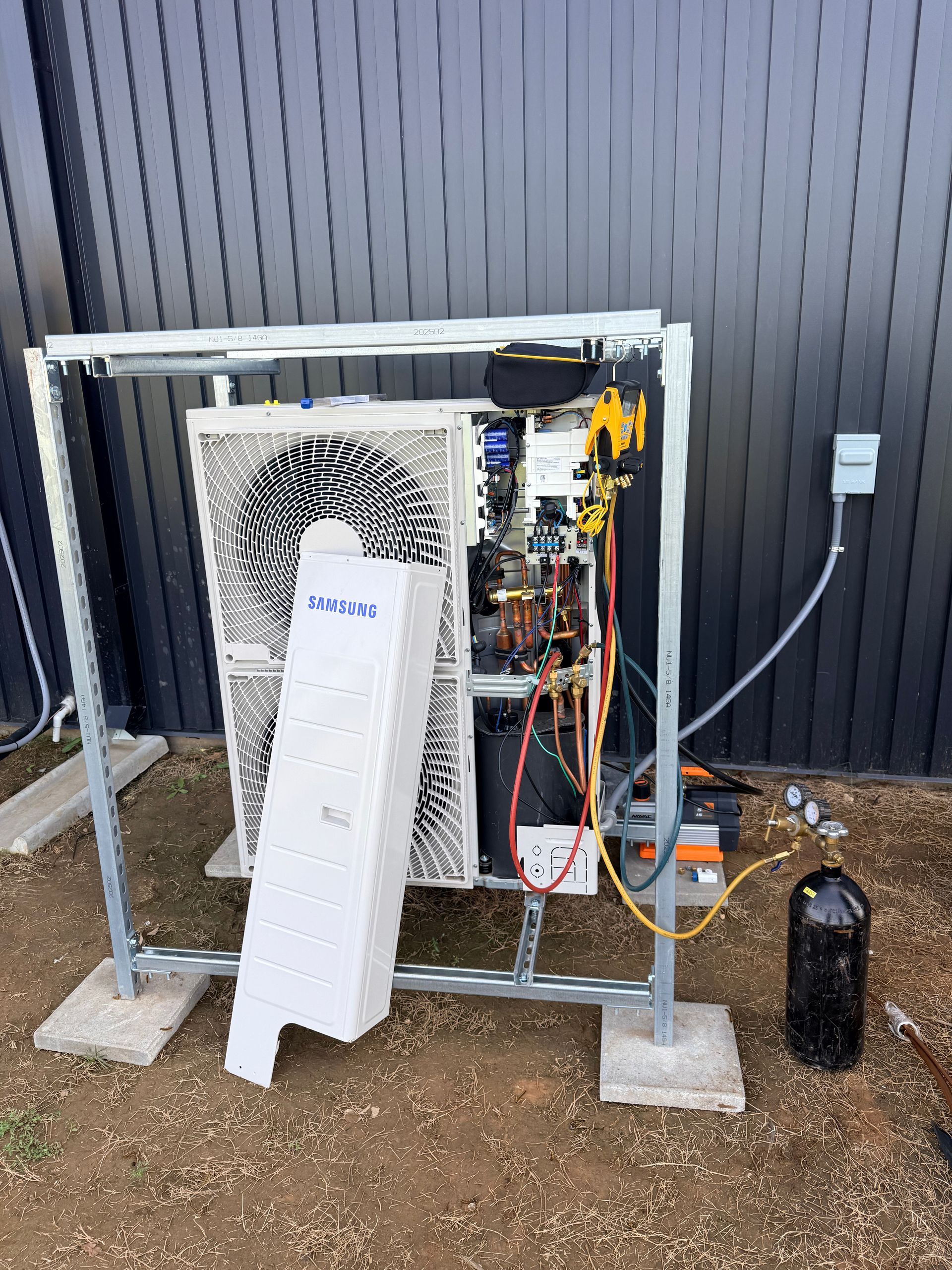 Outdoor HVAC unit mounted on a metal frame beside a building wall, with pipes and cables visible.