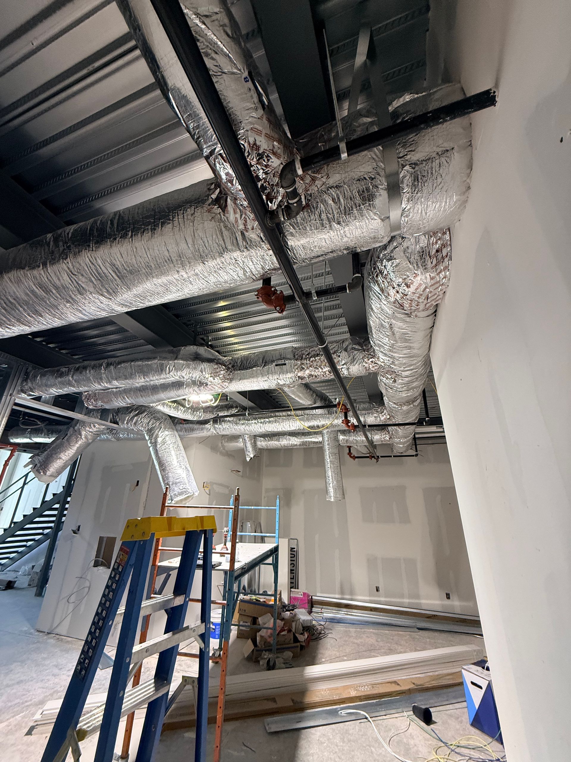Industrial ceiling with exposed ductwork and pipes above a room under construction, with a blue ladder below.