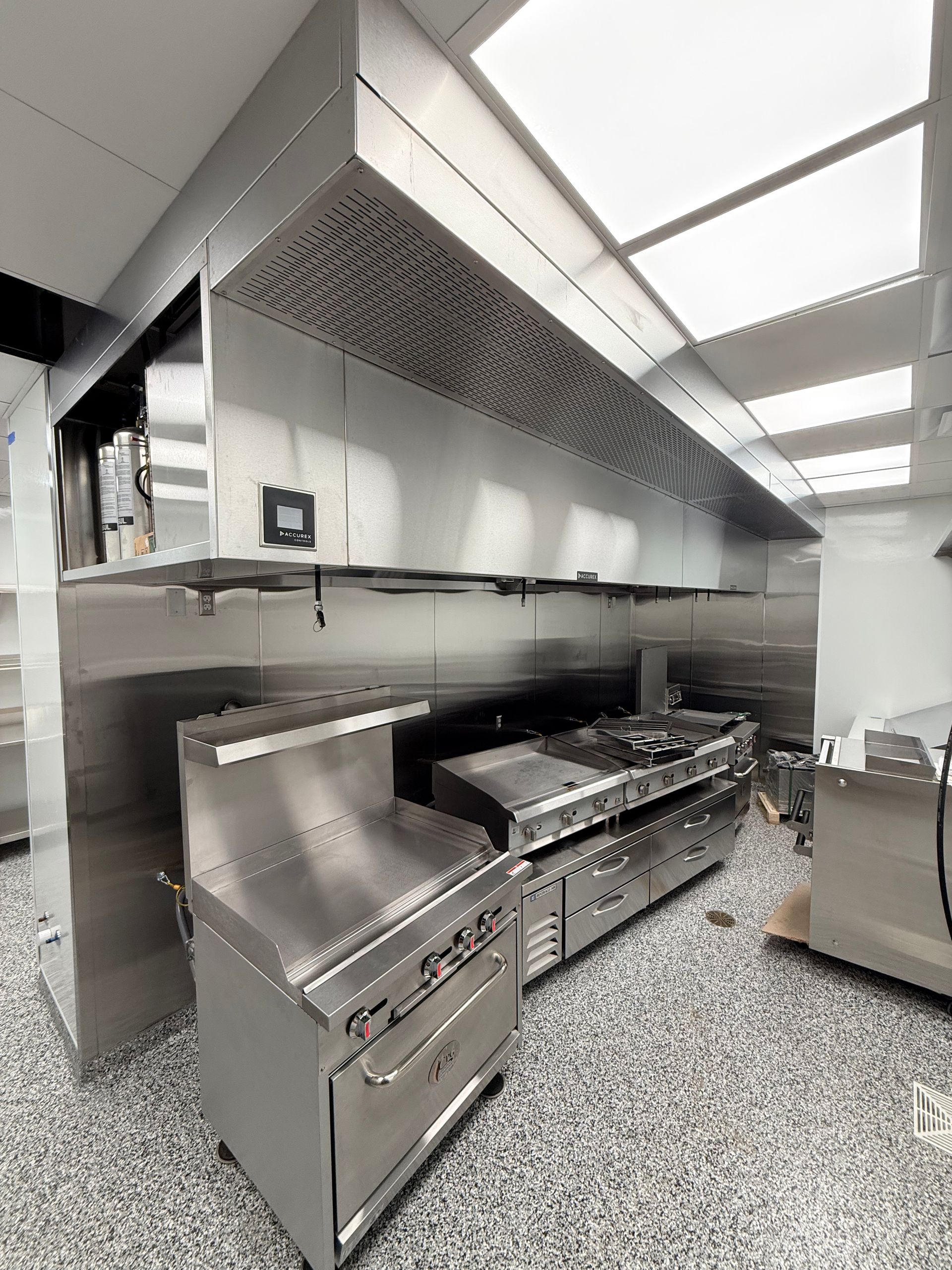 Stainless steel commercial kitchen with grill, fryer, and ventilation hood under bright ceiling lights
