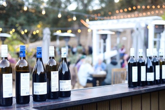 A row of wine bottles sits on a bar counter in front of an outdoor gathering with blurred string lights in the background.