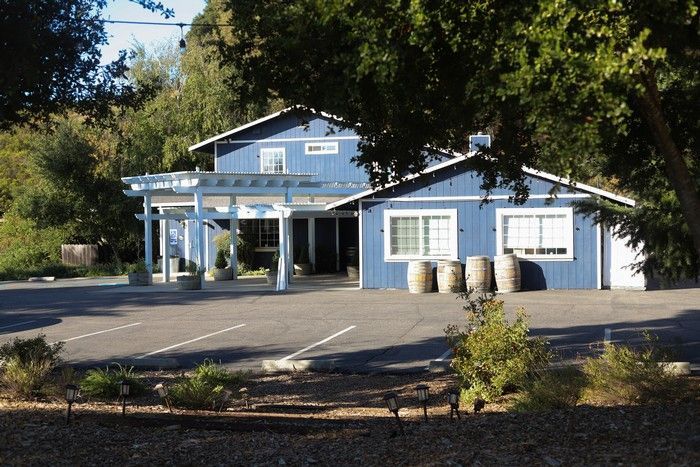 A blue, two-story building with a pergola, parking lot, and barrels in front, surrounded by trees on a sunny day.