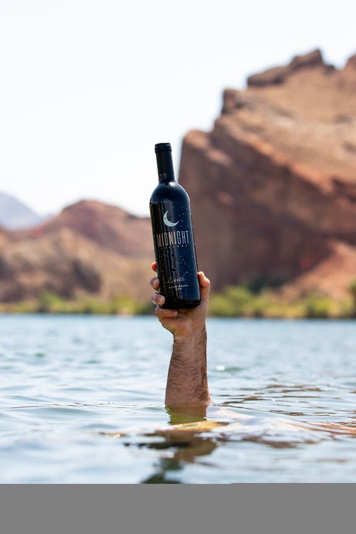 A person’s arm emerges from a lake, holding a bottle of wine aloft against a backdrop of desert mountains.