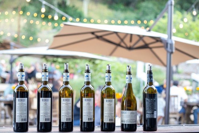Seven wine bottles stand in a row on a wooden surface at an outdoor event with umbrellas and fairy lights in the background.