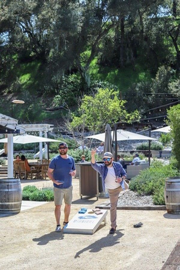 Two people play cornhole on a sunny patio at a winery, with one person in mid-throw and the other observing.