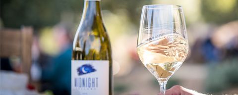 A bottle of Midnight wine and a glass of white wine held by a person outdoors in a soft-focus setting.