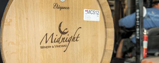 A wooden wine barrel featuring the Midnight Winery & Vineyard logo, with a person operating machinery in the background.