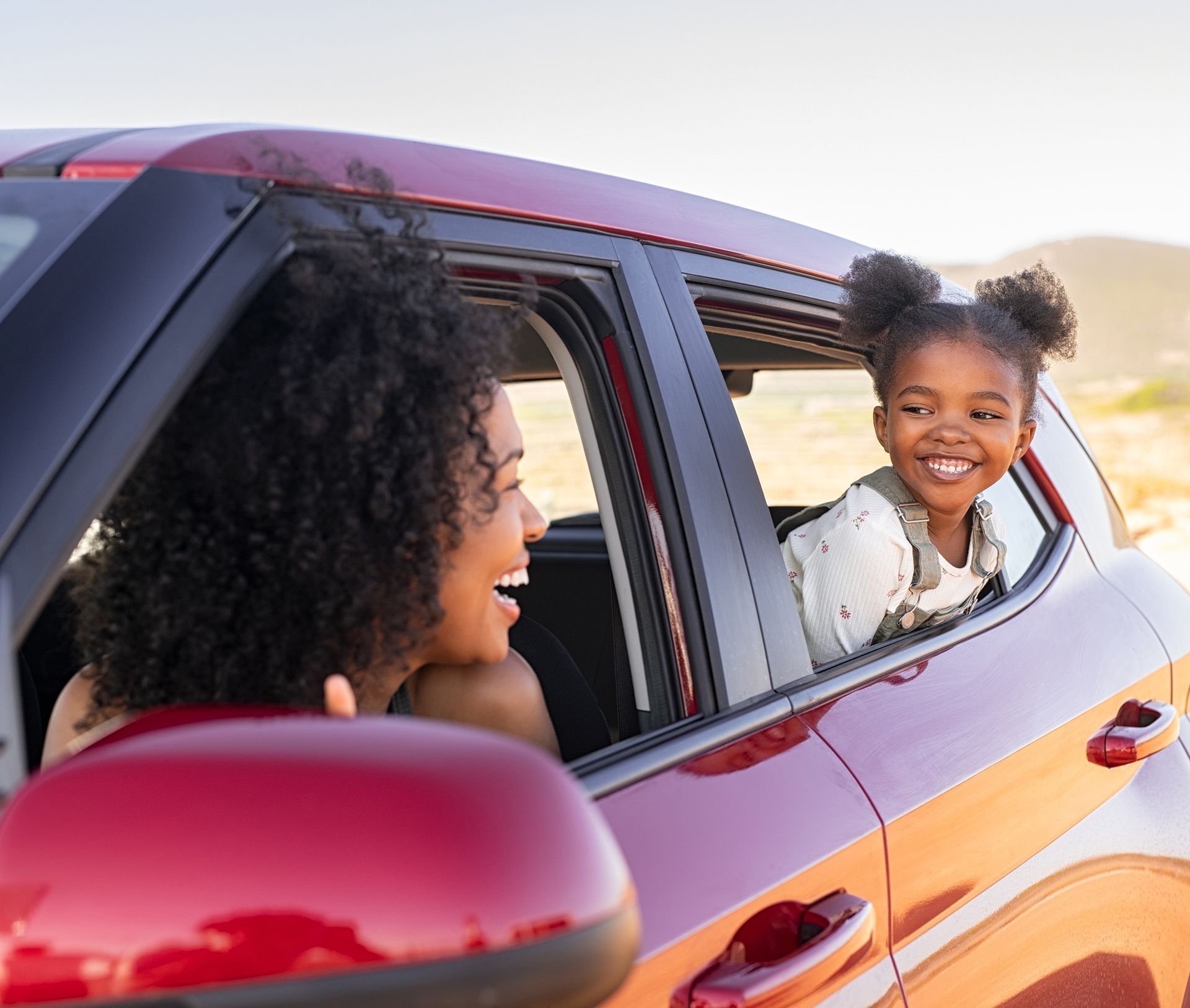 Woman and child in a red car, smiling at each other. Child leaning out the window in a sunny outdoor setting.