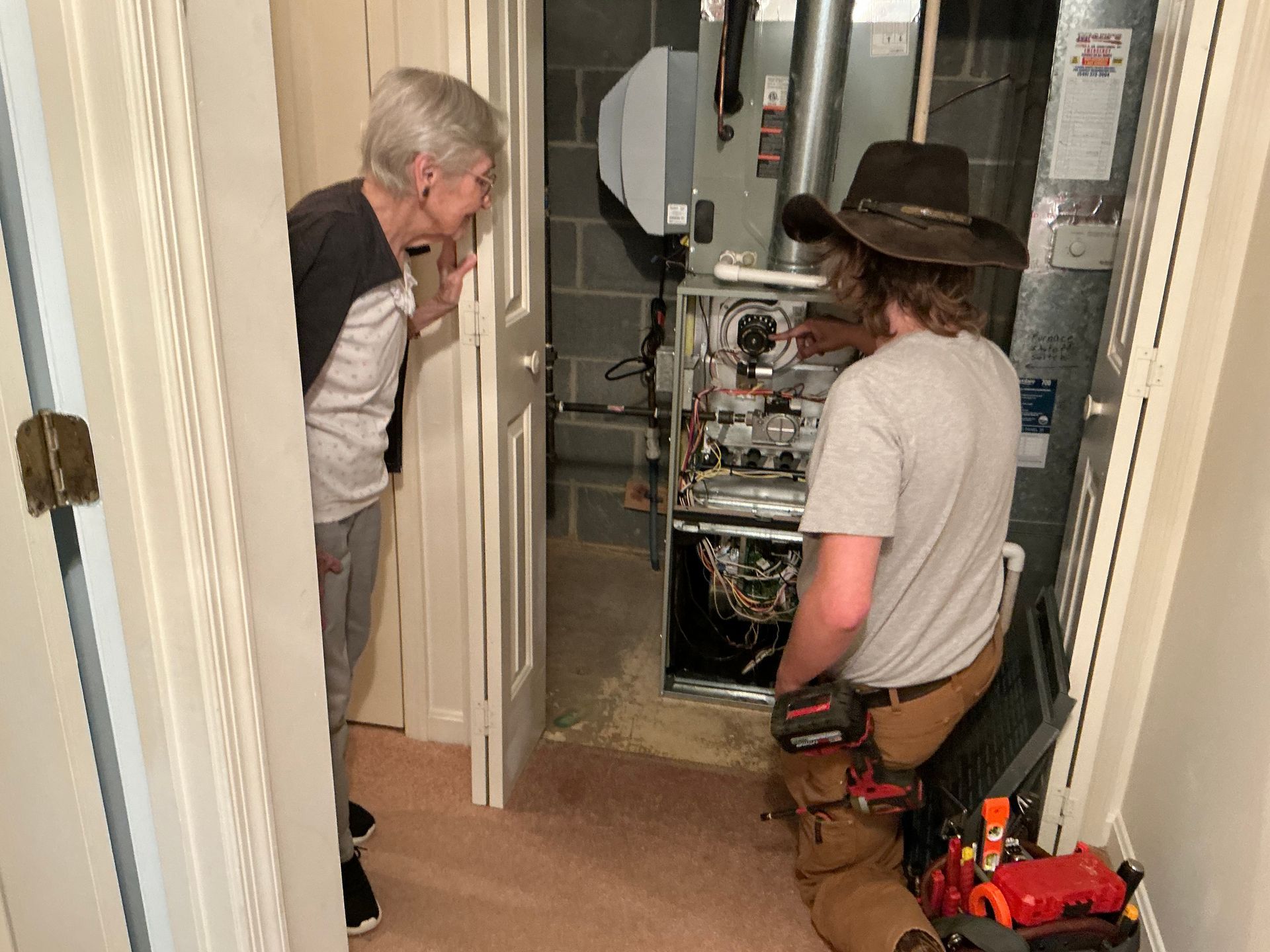 A man in a cowboy hat is working on a furnace while a woman looks on.