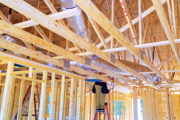 A man is standing on a ladder in the ceiling of a house under construction.