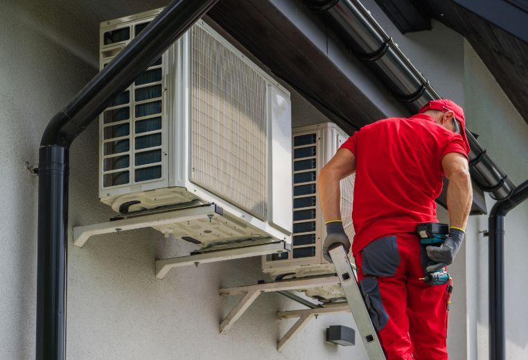 A man is standing on a ladder fixing air conditioners on the side of a building.