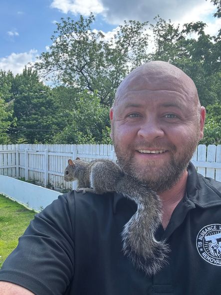 Man smiles with a squirrel on his shoulder outside near a white fence and green trees.