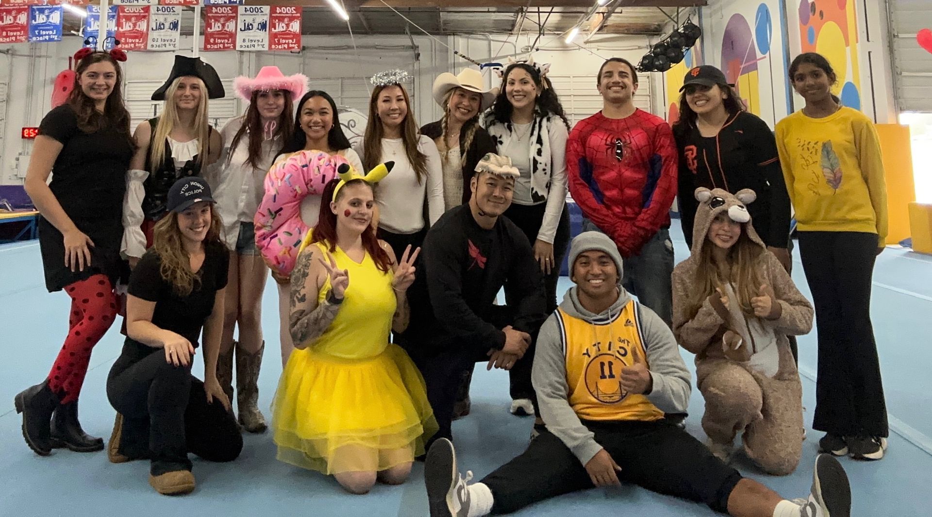 Group of people in costumes posing indoors near a gymnastics area; some are kneeling or sitting.