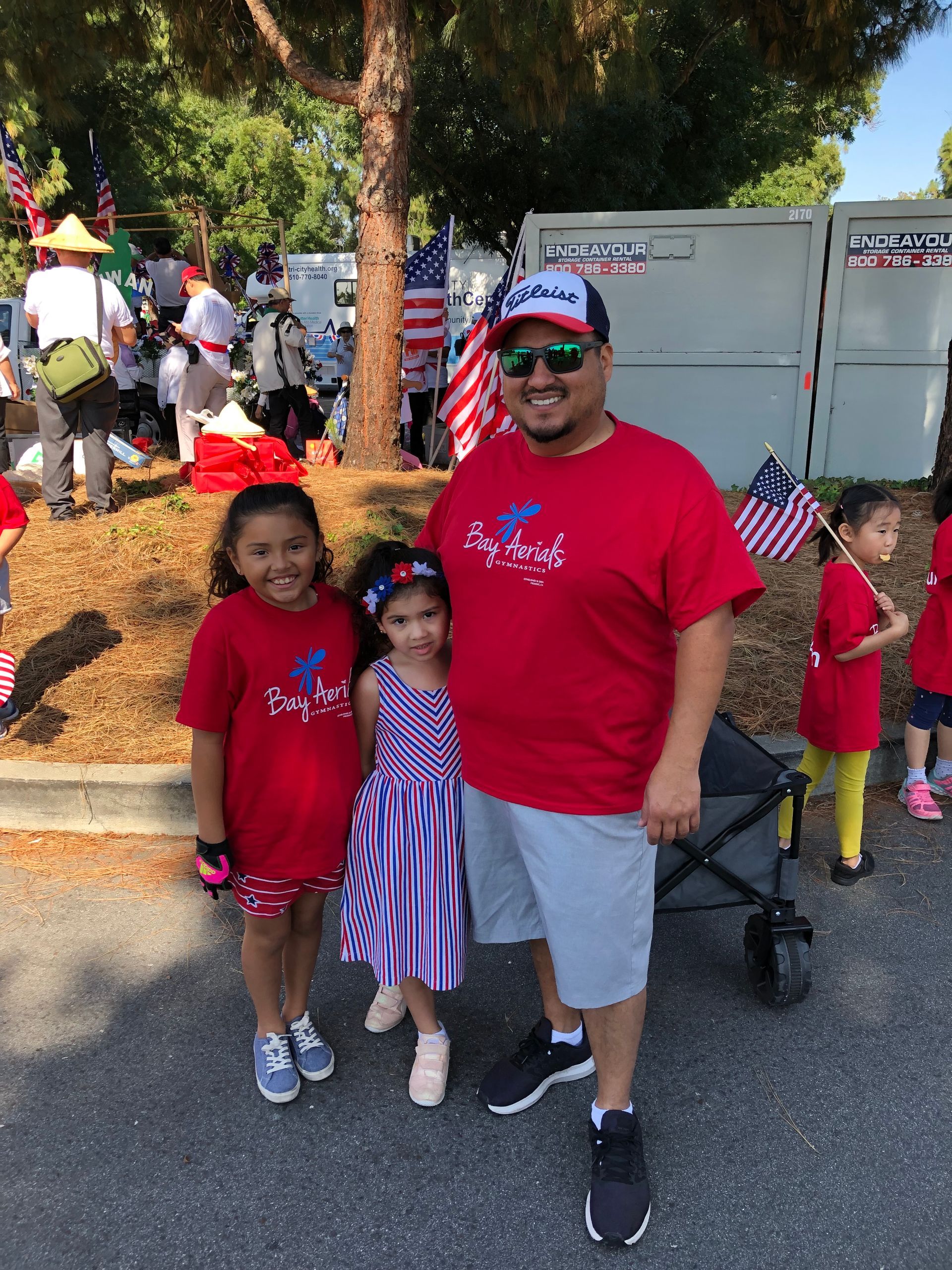 Man and two girls wearing red shirts, outside, possibly at a parade with American flags.