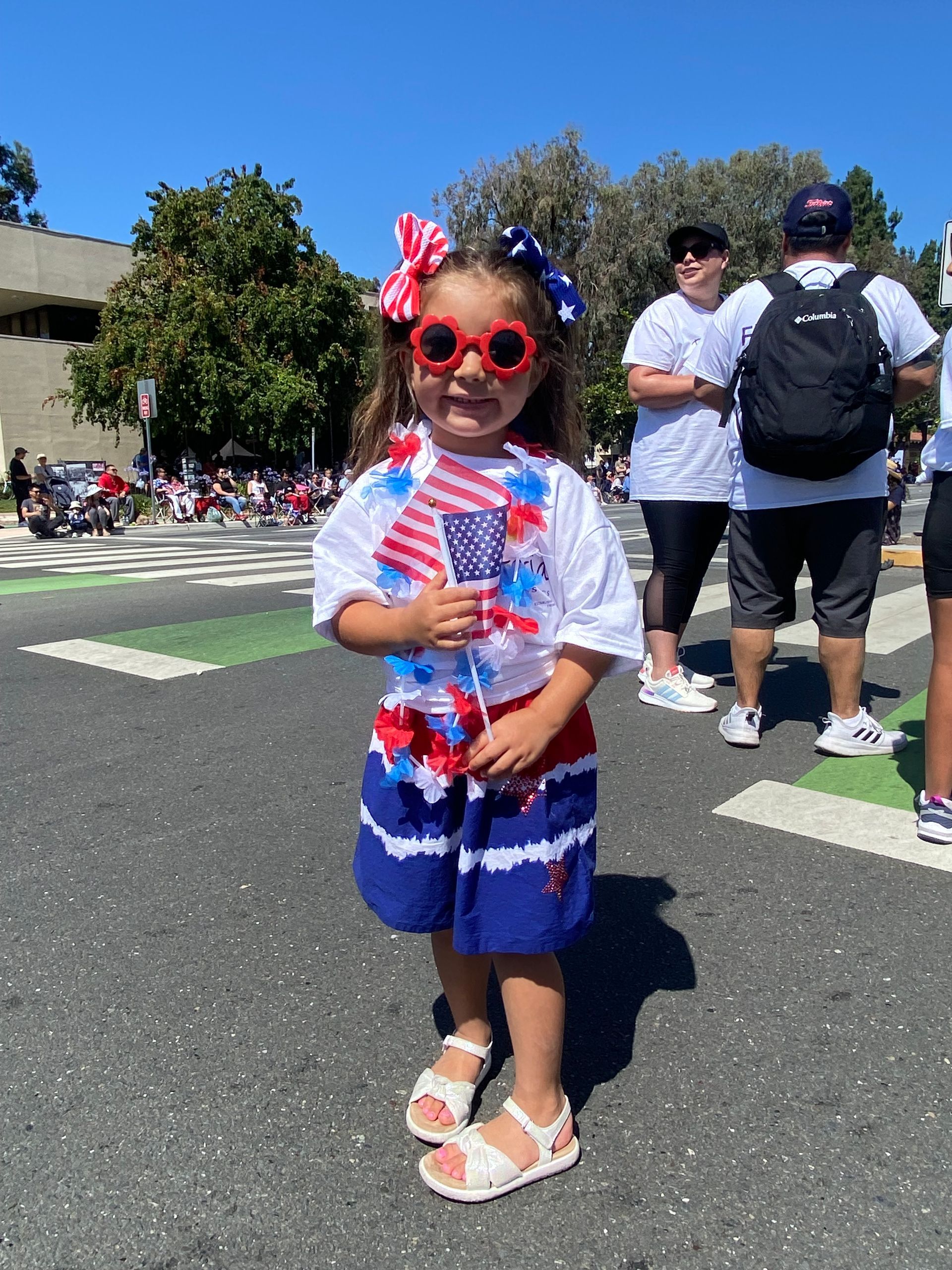 Young child in red, white, and blue patriotic outfit, holding American flag, wearing sunglasses, in a street parade.