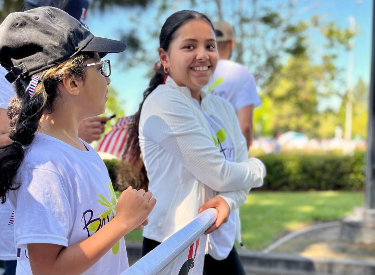 Two people in white shirts smiling, possibly at an event outside. One is wearing a cap.