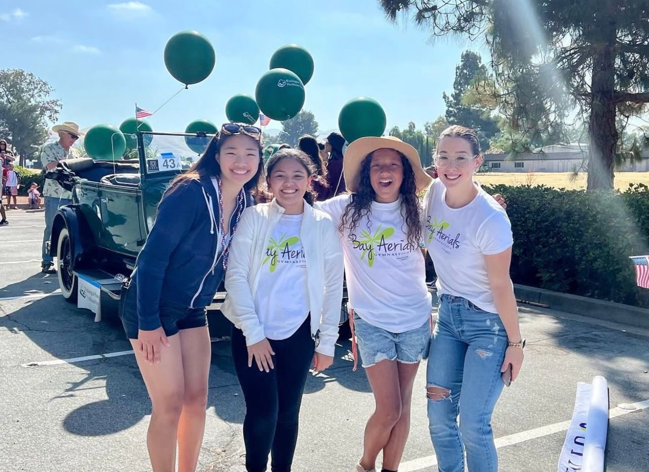 Four smiling people pose in front of a vintage car decorated with balloons. Sunny outdoor setting.