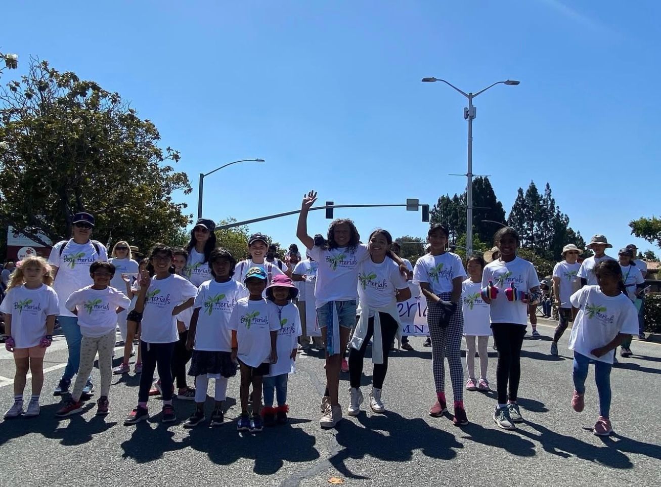 Group of children in matching white shirts walking down a street on a sunny day.