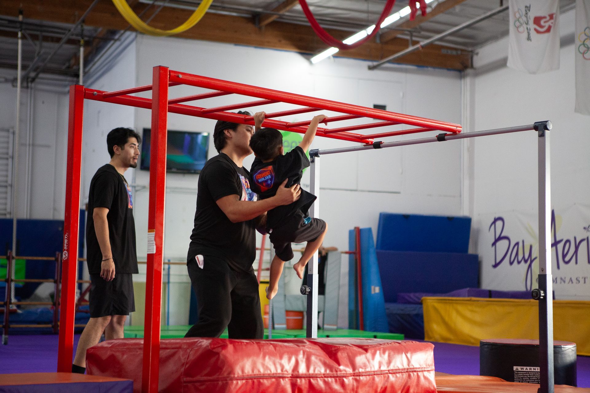 Child climbing a red horizontal ladder with the help of an instructor in a gym; another person watches.